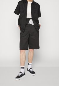 Zwart overhemd met korte mouwen en knoopsluiting, voorzien van twee borstzakken, gecombineerd met zwarte knielange shorts. Accessoires omvatten witte sokken met zwarte strepen en zwarte sneakers.