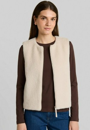 Giovane donna che indossa un gilet in pile color crema sopra una camicia marrone a maniche lunghe e jeans blu, in piedi davanti a uno sfondo grigio uniforme.