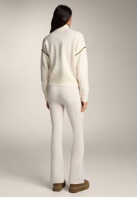 Džemper krem boje, pleteni s visokim ovratnikom i detaljima u smeđem šavu, uparen s uskim svijetlosmeđim hlačama i smeđim platformskim cipelama.