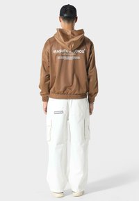 Bruine hoody met witte tekst op de achterkant, gecombineerd met losse witte cargo broeken met grote zakken en een zwarte pet.