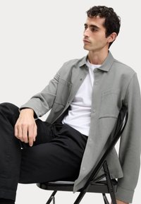 Camisa de un tejido gris con botones al frente, dos bolsillos en el pecho y un cuello extendido, combinada con pantalones negros, sentado en una silla plegable negra.