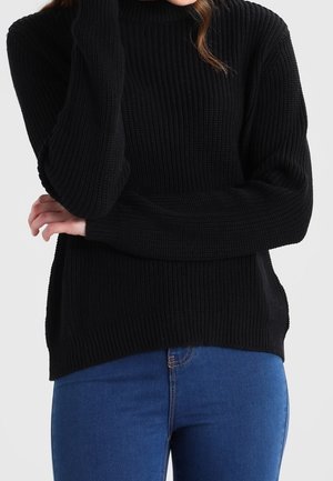 Pull-over en tricot noir avec un col haut, présentant une texture côtelée, des manches longues et une légère fente sur le côté au niveau de l'ourlet.