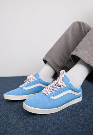 Person trägt hellblaue Sneaker mit weißen Sohlen und gemusterten Schnürsenkeln, weiße Socken und graue Hose, sitzt auf einem blau Teppichboden.