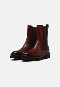 Une paire de bottes en cuir marron mi-mollet avec des panneaux latéraux élastiques noirs, des semelles épaisses en caoutchouc noir et des languettes de tirage sur un fond blanc.