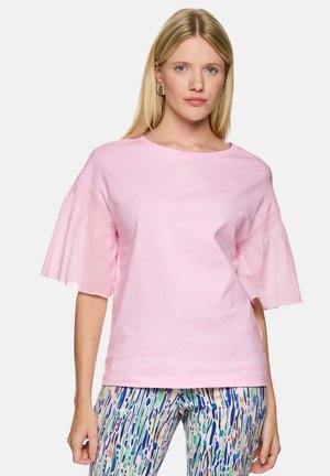 Woman wearing a light pink top with wide, textured half sleeves and multicolored patterned pants against a plain background.