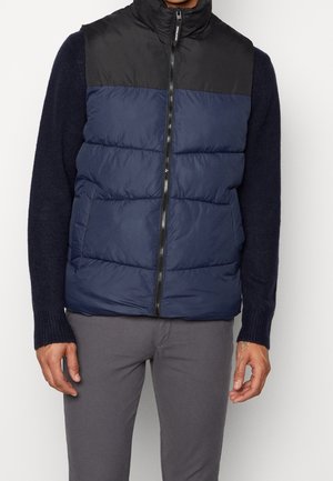 Man wearing a navy and black sleeveless puffer vest over a navy sweater and gray pants, standing against a plain background.