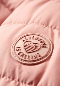 Tessuto di giacca a vento rosa con una patch circolare. La patch presenta il testo in rilievo "L'AVVENTURA STA CHIAMANDO" e un design a cuore con strisce.