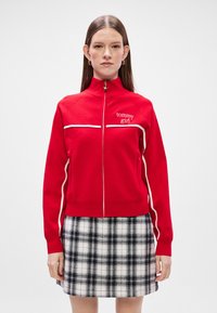 Chaqueta roja con cremallera y acentos blancos, con un cuello alto y el logo "tommy girl". Combinada con una falda de cuadros en blanco y negro.