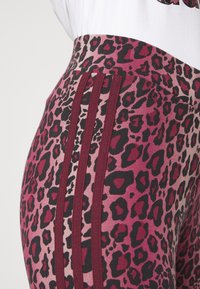 Close-up of pink and black leopard print leggings with dark red three-stripe side detailing and a white top partially visible.