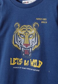 Kék pamut póló, amelyen egy sárga és fekete tigrisfej grafika látható, a következő szövegekkel: "LEGYEN KEMÉNY" és "HEVES HANGULAT" sárga és fehér színben.