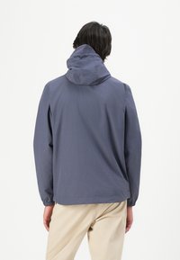 Homme portant une veste à capuche bleu marine et un pantalon beige, se tenant de dos face à l'appareil photo sur un fond uni.