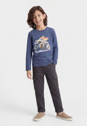 Garçon souriant aux cheveux bruns mi-longs portant une chemise à manches longues bleue avec un motif de monster truck en flammes, un pantalon cargo foncé et des chaussures beige à enfiler.