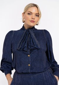 Denimbluse mit gepufften Ärmeln, großem Schleifenband am Hals, goldenen Knopfverschlüssen und kontrastierenden Nähten in Dunkelblau.