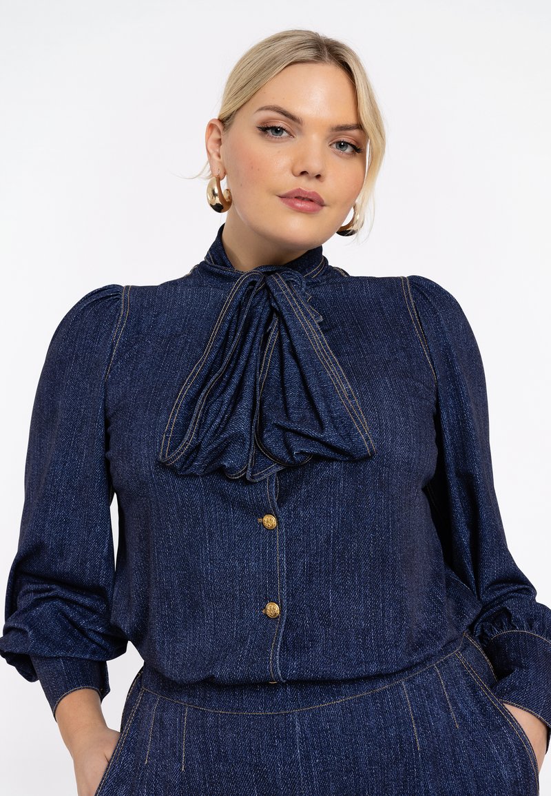 Denimbluse mit gepufften Ärmeln, großem Schleifenband am Hals, goldenen Knopfverschlüssen und kontrastierenden Nähten in Dunkelblau.