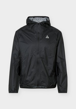 Geacă neagră ușoară cu glugă, fermoar frontal complet, manșete elastice, buzunare laterale și un mic logo triunghiular alb pe piept.