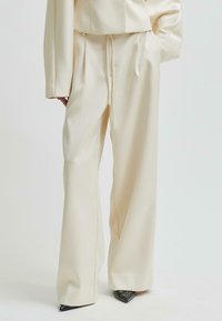 Vidbenade krämfärgade byxor med frontveck och dragsko i midjan, som kombineras med spetsiga svarta skor och en matchande krämfärgad jacka.