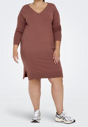 Vrouw draagt een bruine lange mouw gebreide jurk met zijsplitten en witte sportschoenen, staand tegen een effen lichte achtergrond.