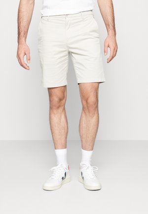 Mand iført lysebeige shorts, hvide sokker og hvide sneakers, der står mod en ensfarvet lys baggrund.