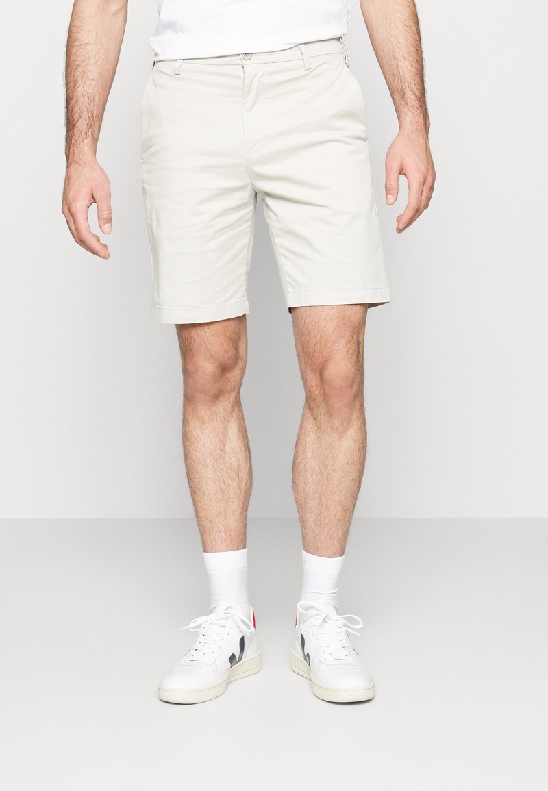 Homme portant un short beige clair, des chaussettes blanches et des baskets blanches, debout devant un fond clair uni.