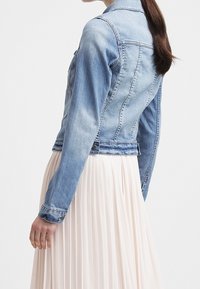 Ljusblå denimjacka med figursydd design, klassisk krage och knäppt fram. Har långa ärmar och en kort modell. Rynkad creamfärgad kjol syns.