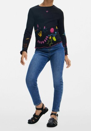 Dziecko ubrane w czarną koszulkę z długim rękawem z kolorowymi nadrukami gwiazdek, niebieskie obcisłe dżinsy i czarne sandały z paskami, stojące na jednolitym tle.