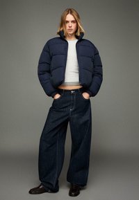 Tmavě modrá prošívaná bunda s vysokým límcem, šedý krátký top, široké tmavě denimové džíny a černé mokasíny, stojící na neutrálním pozadí.