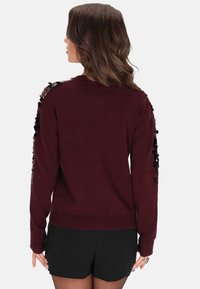 Donna con capelli castano ondulati che indossa un maglione rosso scuro con paillettes nere sulle maniche e pantaloncini neri, in piedi di spalle su uno sfondo bianco.