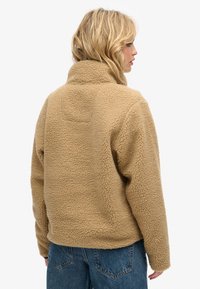 Beige fleece-trøje med høj krave og tekstureret overflade. Har en afslappet pasform og er kombineret med blå jeans.