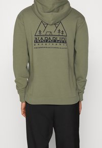 Felpa verde oliva con design geometrico di montagne e alberi neri, testo "NAPAPIJRI geographic", indossata da una persona di spalle, pantaloni neri visibili.