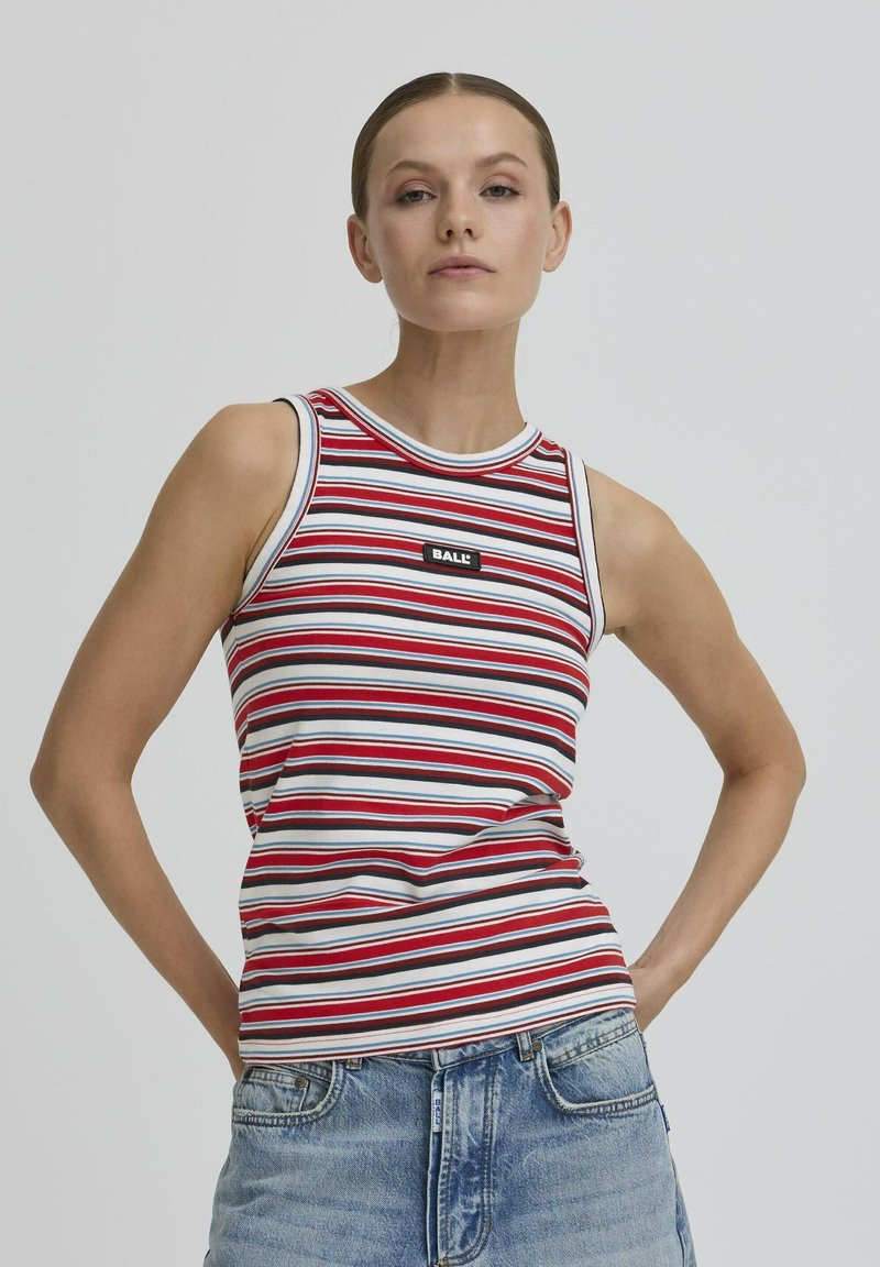 Femme les mains sur les hanches, portant un top sans manches rayé rouge, blanc et bleu, ainsi que un jean en denim bleu clair, devant un fond uni.