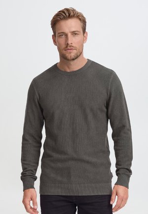 Homme aux cheveux blonds courts et à la barbe portant un pull texturé à manches longues gris foncé et un pantalon noir, debout devant un fond clair uni.