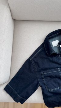 Čierna denimová košeľa s golierom, vreckom a kontrastným zeleným prešívaním, rozložená na svetlom textúrovanom látkovom povrchu.