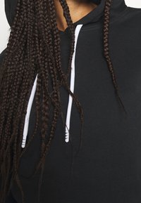 Czarna bluza z kapturem z przednią kieszenią, białymi sznurkami i miękką teksturą. Włosy z długimi, ciemnymi warkoczami częściowo widoczne.