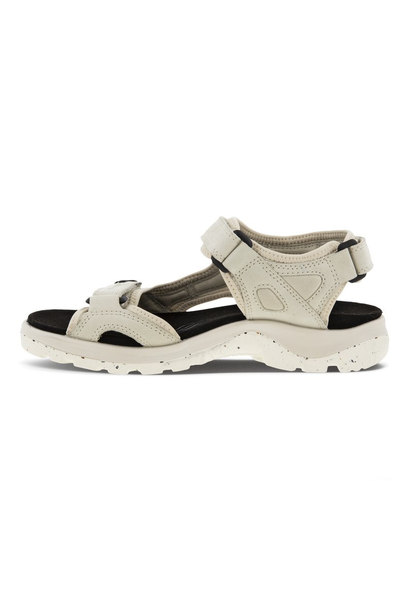 Ljusbeige sandal med två justerbara remmar, svart vadderad innersula, texturerad ovandel och vitprickig sula. Avslappnad utomhusdesign.