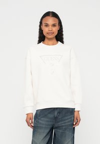 Helles cremefarbenes Sweatshirt mit Rundhalsausschnitt, ausgestattet mit einem dreieckigen silbernen "GUESS"-Logo auf der Brust und gerippten Bündchen. Kombiniert mit Denim.