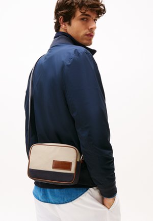 Sac bandoulière avec un devant beige, un fond bleu foncé et un accent en cuir marron. Le tissu a une texture lisse et une forme rectangulaire slim.
