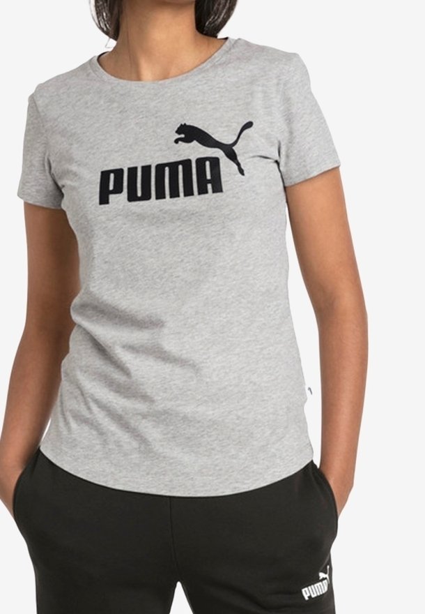Femme portant un t-shirt Puma gris clair avec logo noir et pantalon Puma noir, mains dans les poches, sur fond blanc.