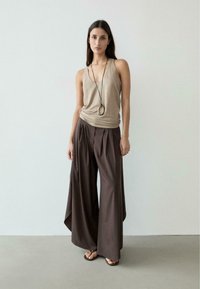 Femme portant un haut beige sans manches, un pantalon large foncé, un long collier pendentif et des sandales noires, se tenant devant un arrière-plan uni clair.