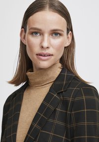 Blazer noir avec un motif à carreaux jaunes sur un col roulé beige. Le blazer possède des revers crantés ; le col roulé a une finition lisse et texturée.