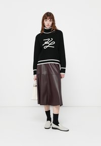 Schwarzer gestrickter Pullover mit weißen Akzenten und Logo, kombiniert mit einem burgunderfarbenen Leder-Midirock, weißen Schuhen und einer beigen Handtasche.