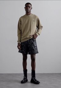 Beiger Pullover mit Logo, schwarze gesteppten Shorts mit Zickzackmuster und glänzende schwarze Stiefeletten, stehend vor einem neutralen Hintergrund.