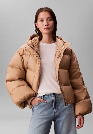 Vrouw die een beige gewatteerde jas met capuchon draagt, een wit shirt en lichtblauwe spijkerbroek tegen een effen grijze achtergrond.