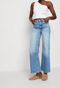 Hellblaue Denim-Jeans mit weit geschnittenem Bein und hoher Taille, ausgestattet mit einem Knopfverschluss und vorderen Taschen. Kombiniert mit einem weißen, kurz geschnittenen Top mit einem Schulterträger.