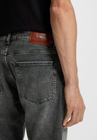 Jeans in denim grigio con un'etichetta in pelle con la scritta "BOSS." Presentano tasche posteriori e una leggera finitura sbiadita. Vestibilità su misura, design minimalista.