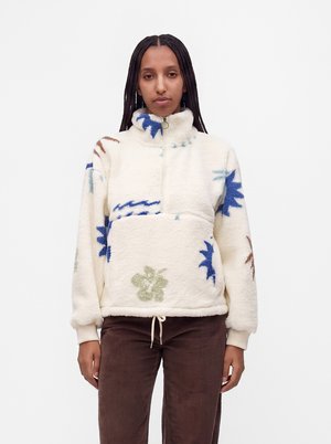 Femme aux longues tresses portant un pull en polaire crème avec des motifs floraux bleus et verts et un pantalon en velours côtelé marron sur fond blanc.