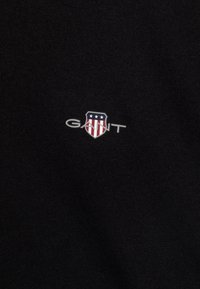 Svart tyg med en liten GANT-logotyp i skölddesign. Skölden visar röda och vita ränder samt stjärnor på en blå bakgrund.
