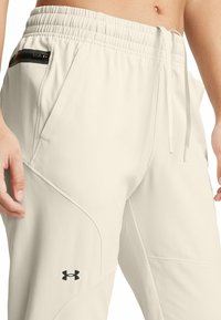 Light beige jogger pants made of soft fabric with a fitted waistband, zippered pocket, and black logo detail on the lower leg.