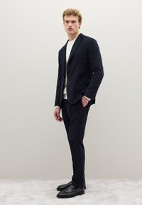 Blazer di velluto a coste blu navy con pantaloni abbinati, caratterizzati da una vestibilità slim e una texture sottile, abbinati a un maglione bianco girocollo e mocassini neri.