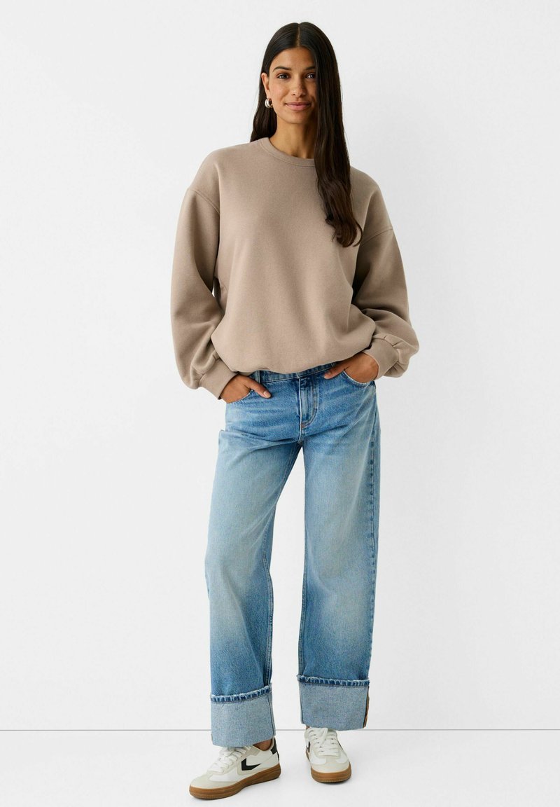 Felpa beige con scollatura rotonda e spalle cadenti, abbinata a jeans a vita alta di colore blu chiaro e con gamba larga, e sneakers bianche.