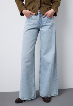 Persona che indossa jeans larghi azzurri, scarpe marroni e una giacca marrone con le mani nelle tasche, in piedi contro un muro grigio semplice.